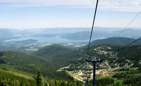 [The support poles and overhead rails of ski lift are on teh right side of the image. There are evergreens all over the ground below except where the resort buildings are located. In the distance are the meanderings of the lake waters.]